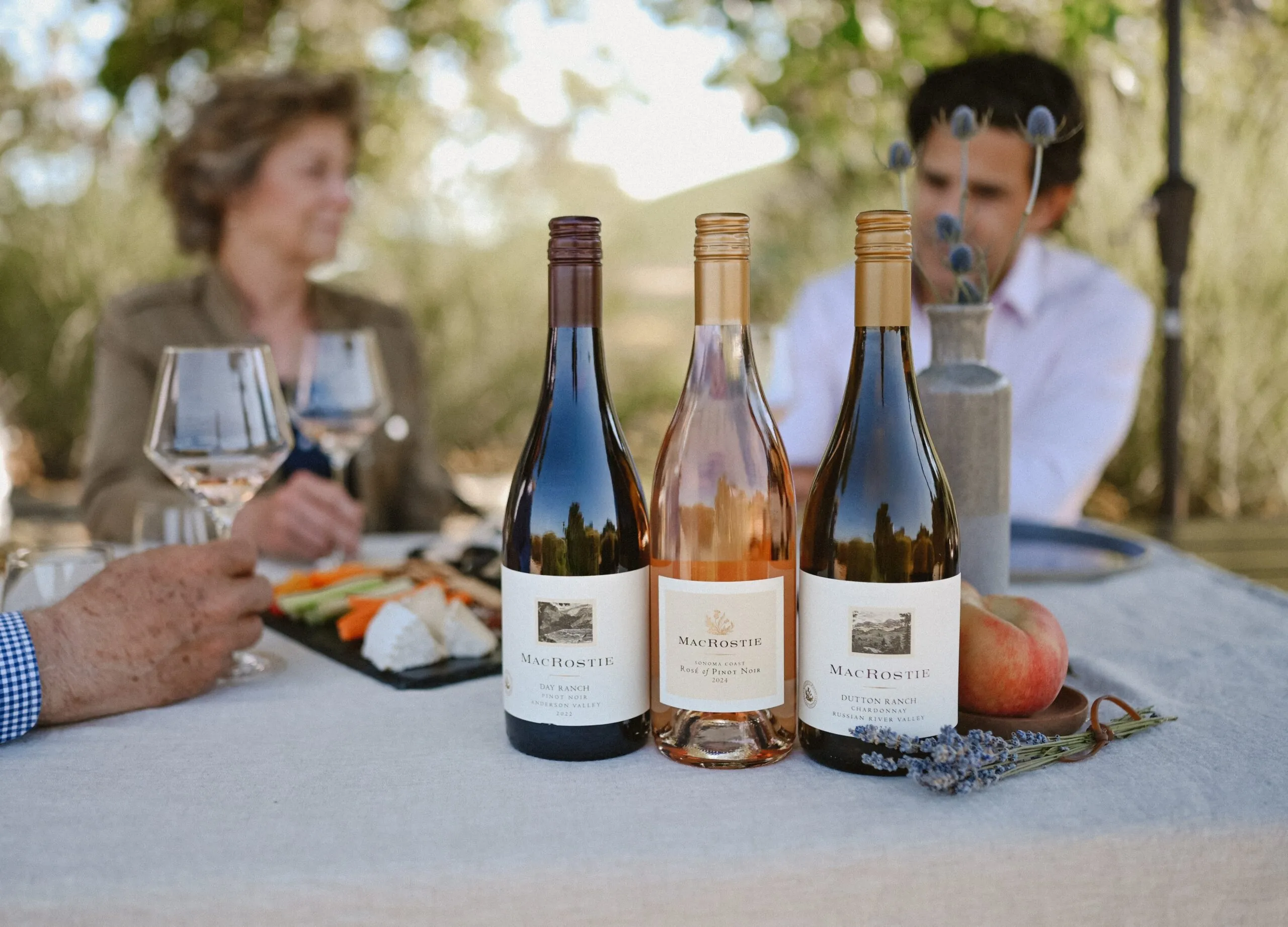 Three bottles of MacRostie Wine on a table with food and people holding glasses of wine