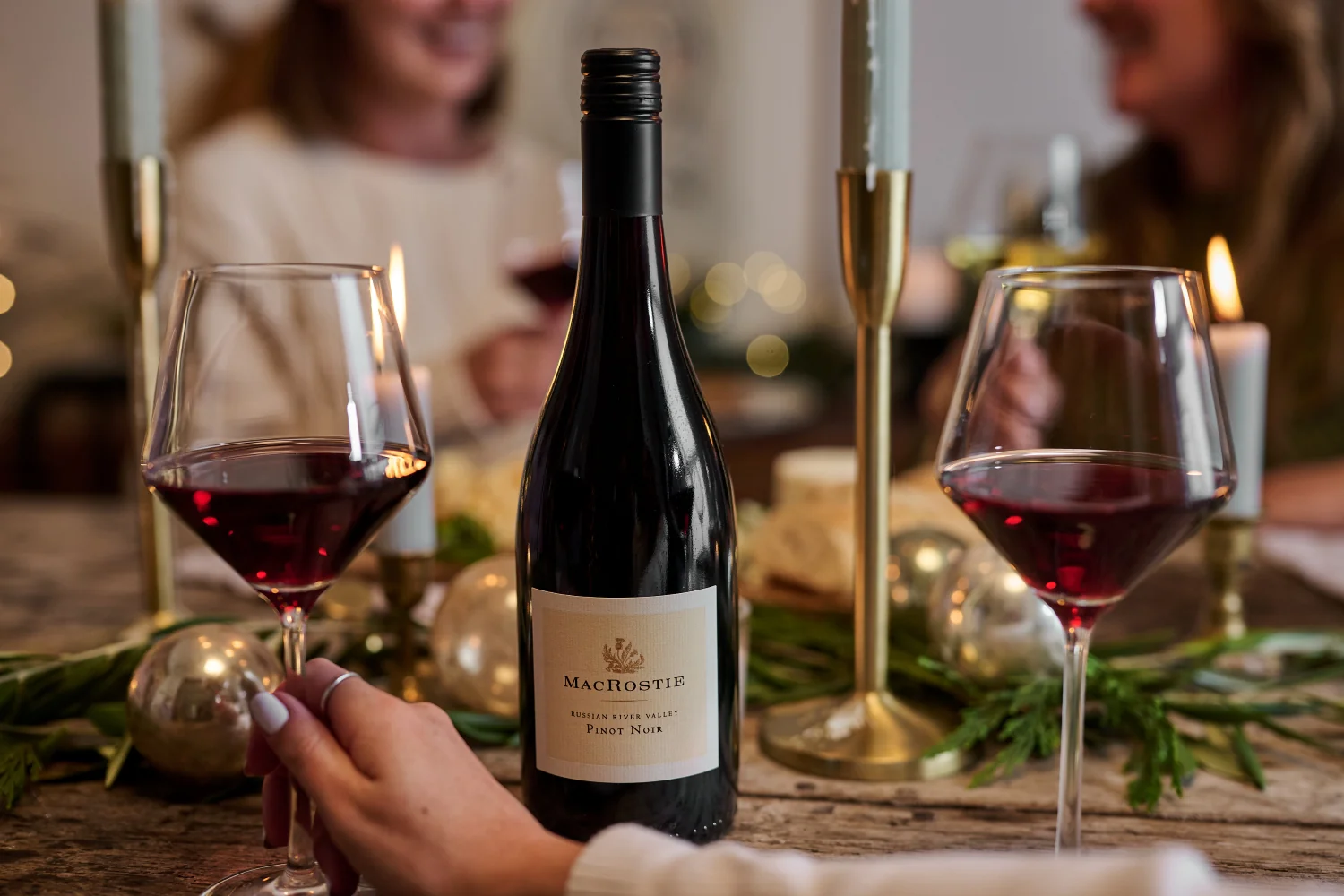 Bottle of MacRostie wine on a table next to a hand holding a glass of wine with people smiling in the background