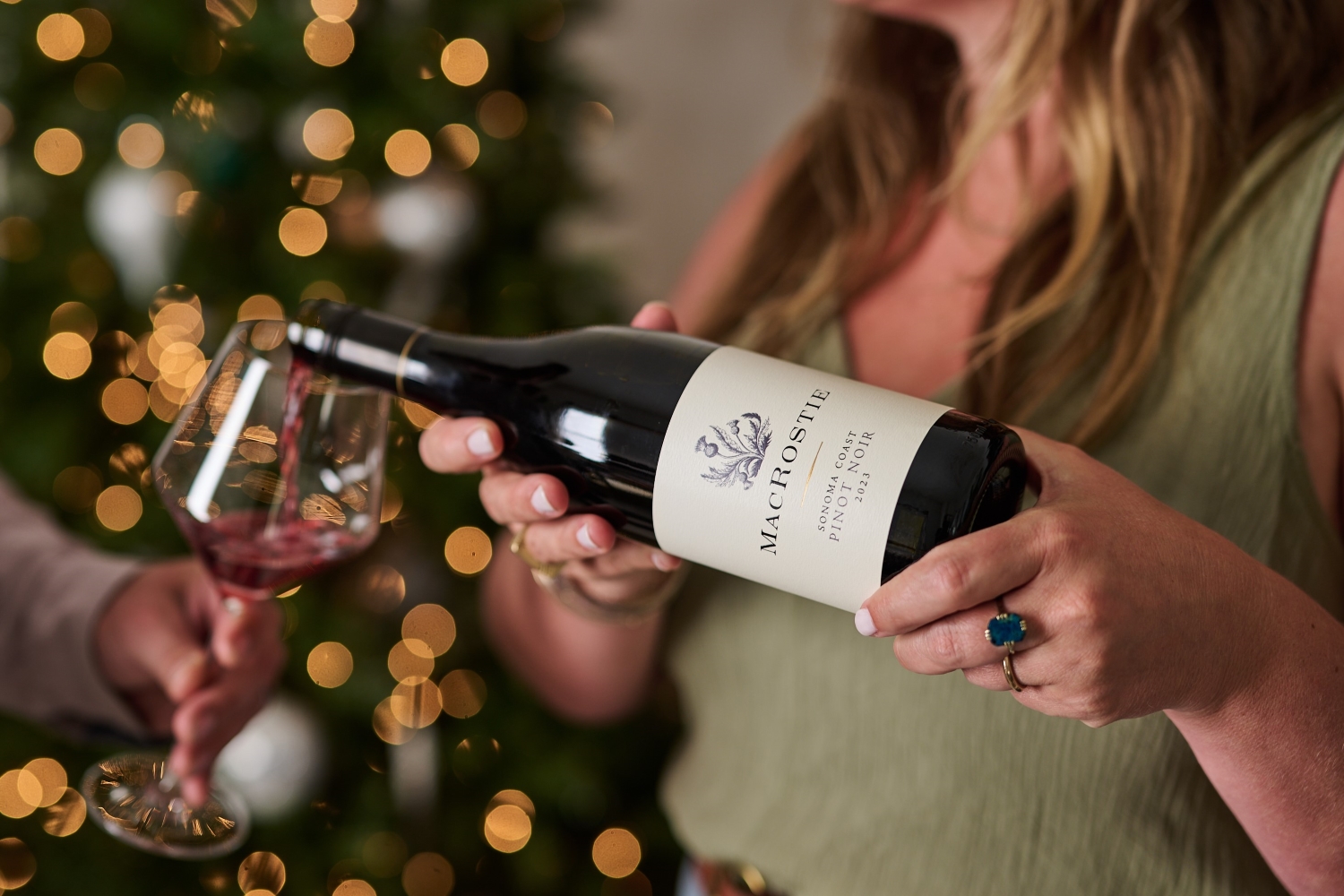 Hands pouring a glass of MacRostie Sonoma Coast Pinot Noir into a glass with a Christmas tree in the background