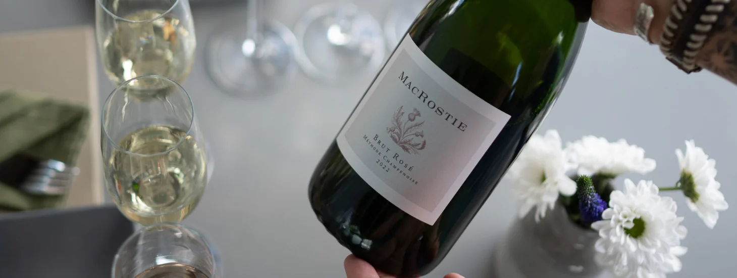 Hands holding a bottle of MacRostie sparkling wine over a table with wine glasses and flowers on it