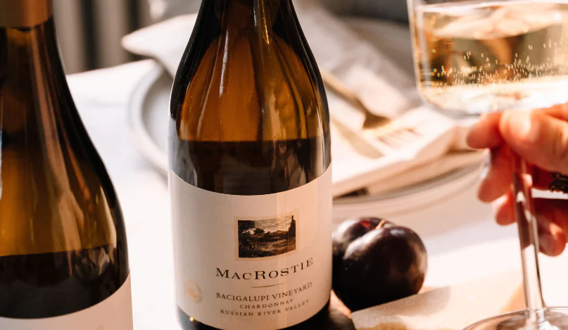 A bottle of MacRostie wine on a table next to food pairings and a hand holding a glass of wine